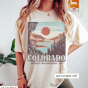 May include: Ivory-colored t-shirt featuring a Colorado landscape design with a red sun, mountains, and a river. The text "COLORADO" and "Olivia's Bachelorette '26" are printed below the graphic. The shirt also has the text "personalize me!"