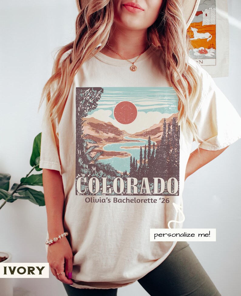 May include: Ivory-colored t-shirt featuring a Colorado landscape design with a red sun, mountains, and a river. The text "COLORADO" and "Olivia's Bachelorette '26" are printed below the graphic. The shirt also has the text "personalize me!"