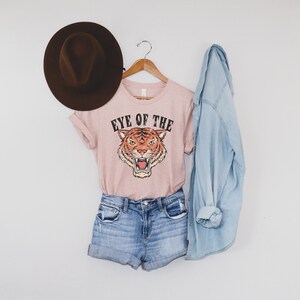 May include: A pink t-shirt with a tiger graphic and the text "Eye of the Tiger". The shirt is paired with denim shorts and a light blue denim jacket.