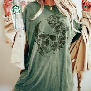 May include: Olive green t-shirt featuring a detailed skull graphic with floral accents. The design is printed on a comfortable, oversized tee. The shirt is a relaxed fit, perfect for casual wear. The image also shows a clear Starbucks tumbler.