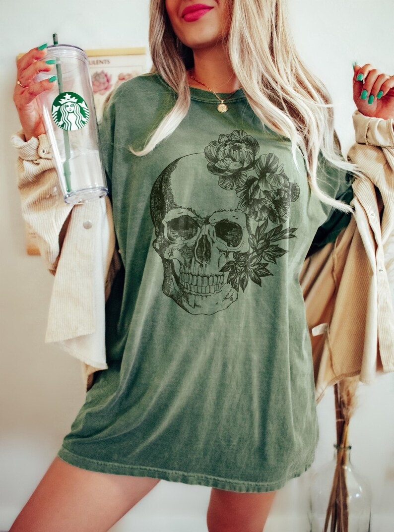 May include: Olive green t-shirt featuring a detailed skull graphic with floral accents. The design is printed on a comfortable, oversized tee. The shirt is a relaxed fit, perfect for casual wear. The image also shows a clear Starbucks tumbler.