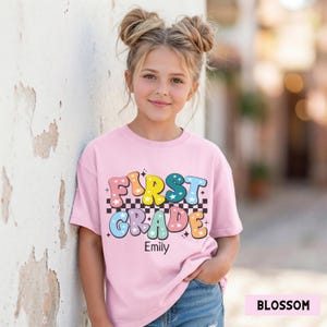 May include: A light pink t-shirt with the words "FIRST GRADE" in colorful, retro-style lettering. The shirt has a checkered pattern and star accents. The name "Emily" is printed below the text.