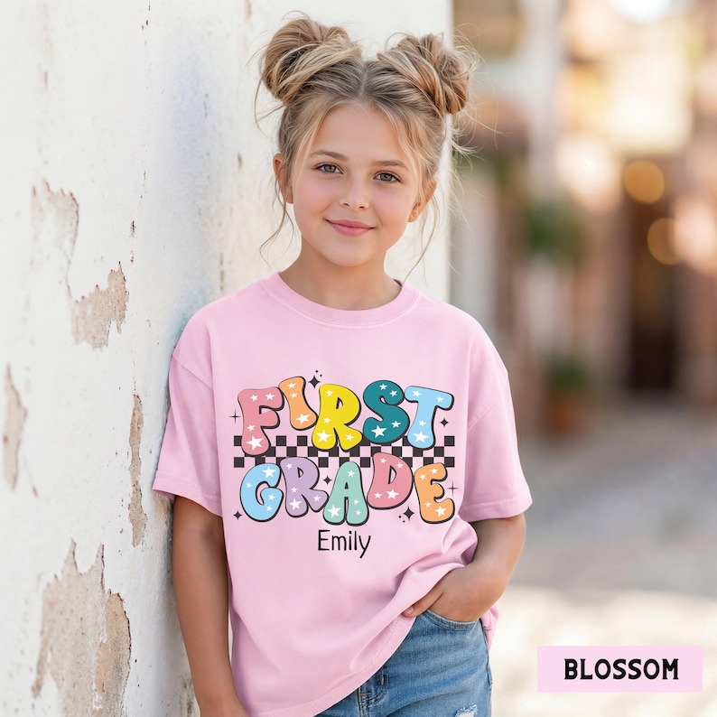May include: A light pink t-shirt with the words "FIRST GRADE" in colorful, retro-style lettering. The shirt has a checkered pattern and star accents. The name "Emily" is printed below the text.
