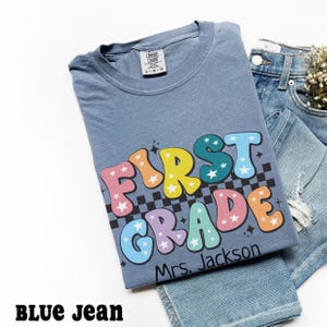 May include: A blue t-shirt with the words "FIRST GRADE" in colorful bubble letters with star accents and a black and white checkered pattern. The name "Mrs. Jackson" is printed below the words. The shirt is paired with blue jeans.
