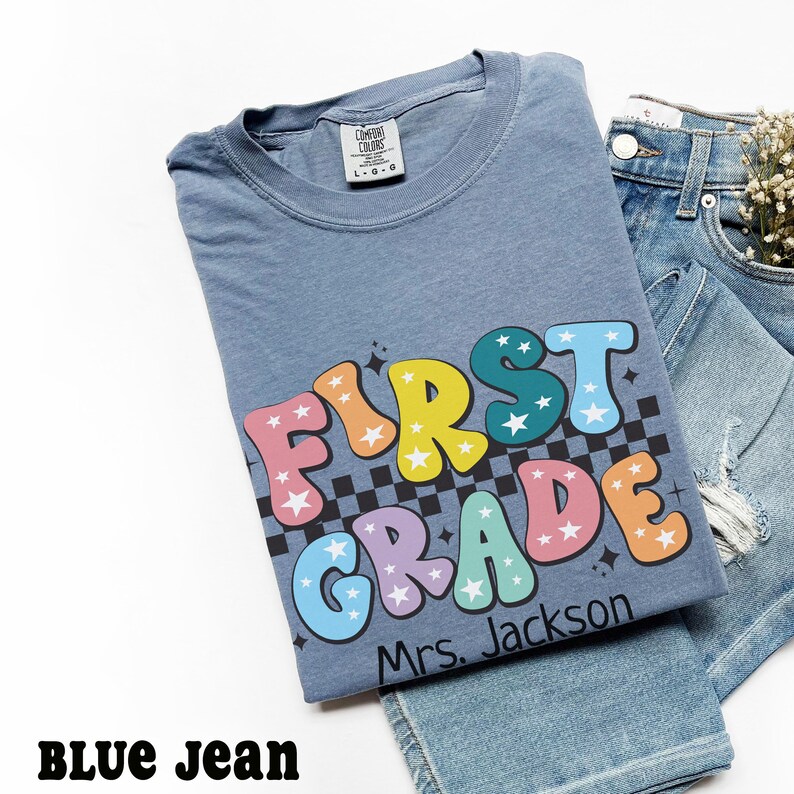 May include: A blue t-shirt with the words "FIRST GRADE" in colorful bubble letters with star accents and a black and white checkered pattern. The name "Mrs. Jackson" is printed below the words. The shirt is paired with blue jeans.