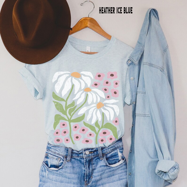K&ouml;nnte beinhalten: Ein hellblaues T-Shirt mit einem Blumendruck aus wei&szlig;en G&auml;nsebl&uuml;mchen und rosa Bl&uuml;ten. Das Shirt h&auml;ngt an einem Kleiderb&uuml;gel und hat den Text "HEATHER ICE BLUE" dar&uuml;ber.
