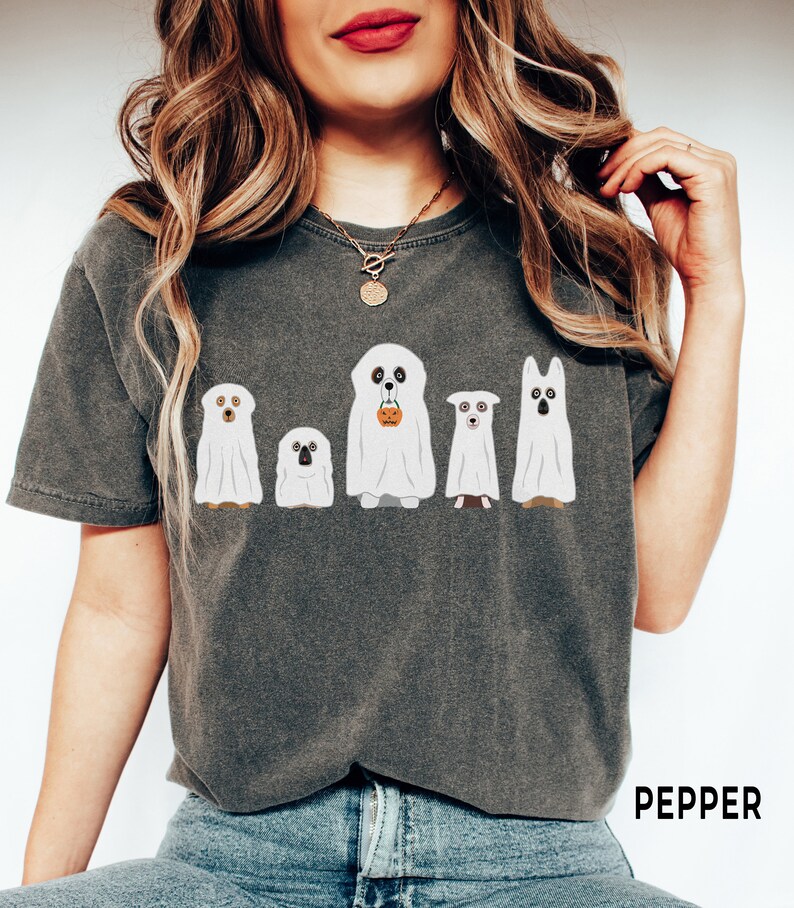 May include: A dark gray t-shirt with a graphic of five dogs dressed as ghosts for Halloween. The dogs are wearing white sheets with faces peeking out. One dog is holding a small orange pumpkin. The text "PEPPER" is printed on the bottom right corner of the shirt.