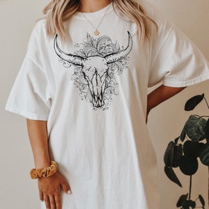 K&ouml;nnte beinhalten: Wei&szlig;es Oversize-T-Shirt mit einem schwarz-wei&szlig;en Grafikdruck eines Bullensch&auml;dels mit floralen Details.