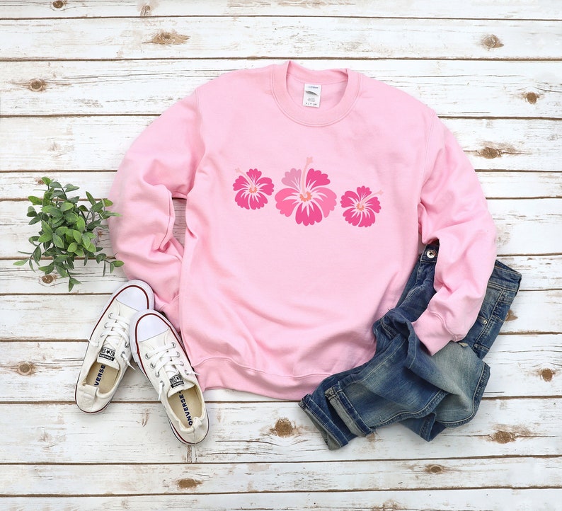 May include: A pink sweatshirt with a row of three pink hibiscus flowers printed on the front. The sweatshirt is laying on a white wooden surface with a pair of white sneakers and blue jeans.