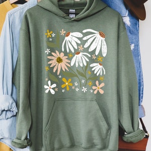 K&ouml;nnte beinhalten: Ein gr&uuml;ner Kapuzenpullover mit einem Blumendruck mit wei&szlig;en, rosa und gelben Blumen. Der Text "MILITARY GREEN" ist unter dem Bild gedruckt.