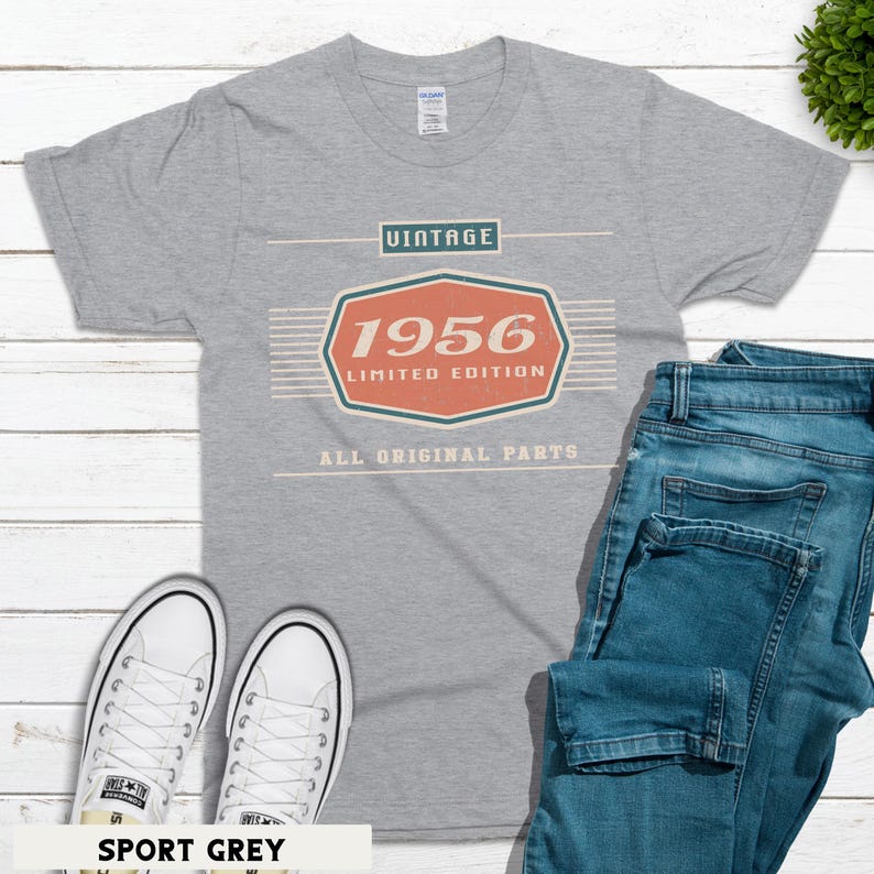 May include: Sport grey t-shirt with the word "VINTAGE" above a red and orange graphic that says "1956 LIMITED EDITION ALL ORIGINAL PARTS". The shirt is paired with blue jeans and white sneakers.