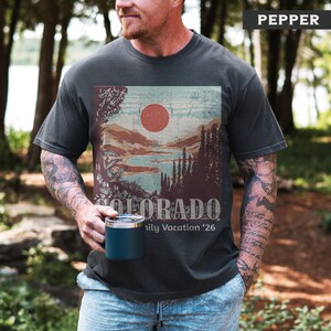 May include: Dark gray t-shirt with a Colorado landscape graphic featuring a red sun, mountains, and trees. The text "COLORADO" and "Family Vacation '26" are printed below the graphic. The shirt is a Pepper color.
