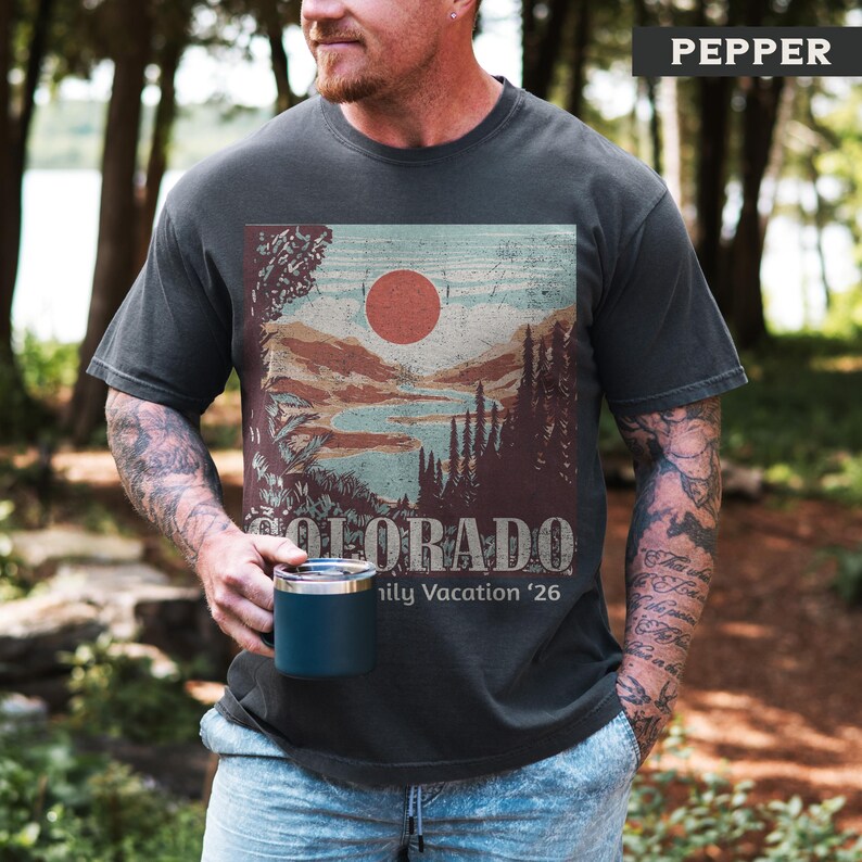 May include: Dark gray t-shirt with a Colorado landscape graphic featuring a red sun, mountains, and trees. The text "COLORADO" and "Family Vacation '26" are printed below the graphic. The shirt is a Pepper color.