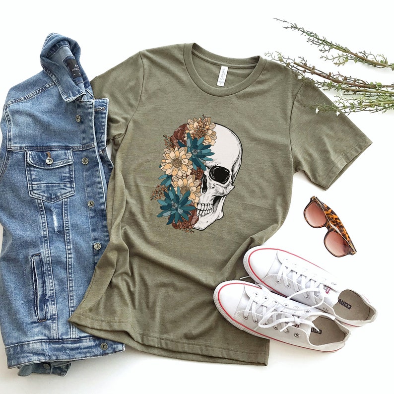 Op de afbeelding: Een groen T-shirt met een wit schedelontwerp. De schedel is half bedekt met kleurrijke bloemen. Het T-shirt ligt op een wit oppervlak met een spijkerjack, witte sneakers en een zonnebril.
