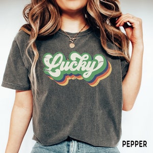 May include: A dark gray t-shirt with a retro-style graphic that says "Lucky" in green letters with a rainbow-colored background.