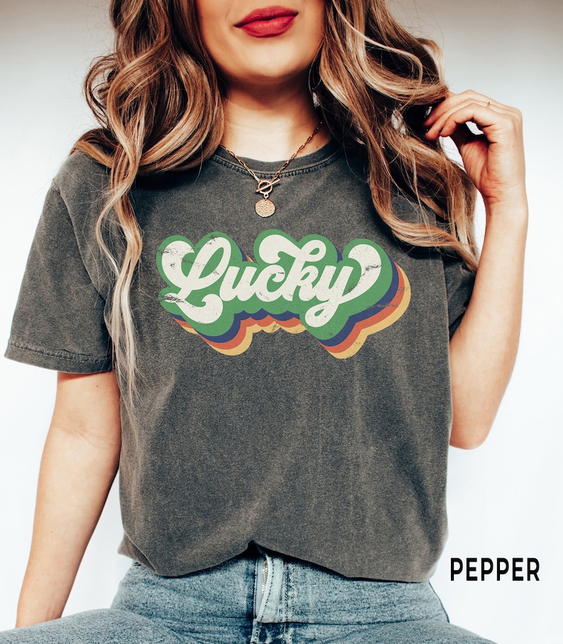 May include: A dark gray t-shirt with a retro-style graphic that says "Lucky" in green letters with a rainbow-colored background.