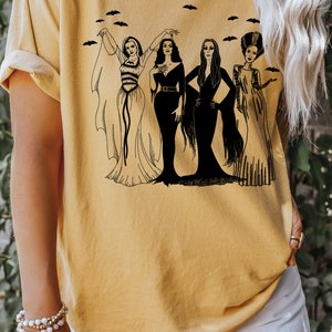 K&ouml;nnte beinhalten: Ein gelbes T-Shirt mit einer schwarzen Grafik von vier Frauen in Vintage-Halloween-Kost&uuml;men. Die Frauen stehen in einer Reihe, und um sie herum fliegen Flederm&auml;use.