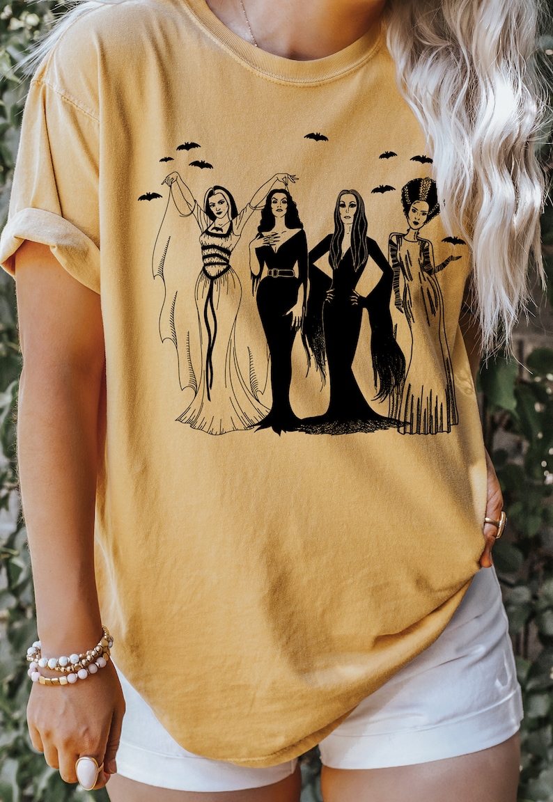 K&ouml;nnte beinhalten: Ein gelbes T-Shirt mit einer schwarzen Grafik von vier Frauen in Vintage-Halloween-Kost&uuml;men. Die Frauen stehen in einer Reihe, und um sie herum fliegen Flederm&auml;use.