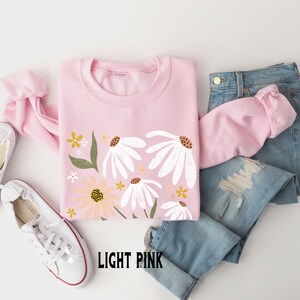 K&ouml;nnte beinhalten: Ein hellrosa Sweatshirt mit einem Blumendruck mit wei&szlig;en G&auml;nsebl&uuml;mchen und gelben Blumen. Der Text "LIGHT PINK" ist unten auf dem Sweatshirt aufgedruckt.