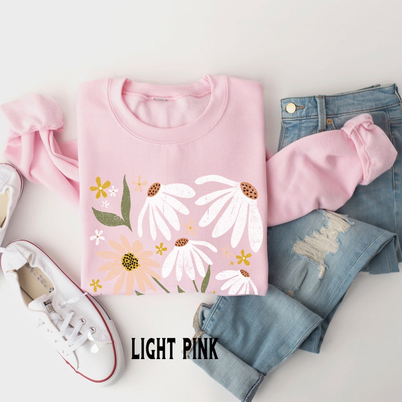 K&ouml;nnte beinhalten: Ein hellrosa Sweatshirt mit einem Blumendruck mit wei&szlig;en G&auml;nsebl&uuml;mchen und gelben Blumen. Der Text "LIGHT PINK" ist unten auf dem Sweatshirt aufgedruckt.