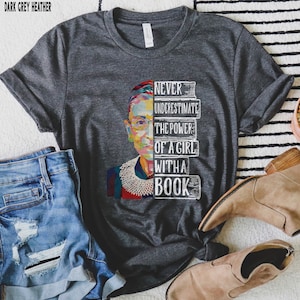 Op de afbeelding: Donkergrijze heather t-shirt met een kleurrijke afbeelding van een vrouwenportret en de tekst "NEVER UNDERESTIMATE THE POWER OF A GIRL WITH A BOOK". Het shirt wordt gecombineerd met een blauwe spijkerbroek en bruine laarzen.