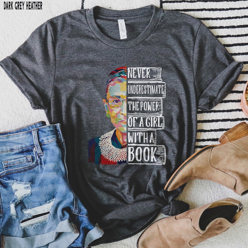 Op de afbeelding: Donkergrijze heather t-shirt met een kleurrijke afbeelding van een vrouwenportret en de tekst "NEVER UNDERESTIMATE THE POWER OF A GIRL WITH A BOOK". Het shirt wordt gecombineerd met een blauwe spijkerbroek en bruine laarzen.