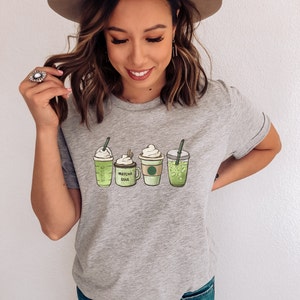 May include: A gray t-shirt with a graphic of four green drinks. The drinks are labeled "Matcha Soul" and "Green Tea".