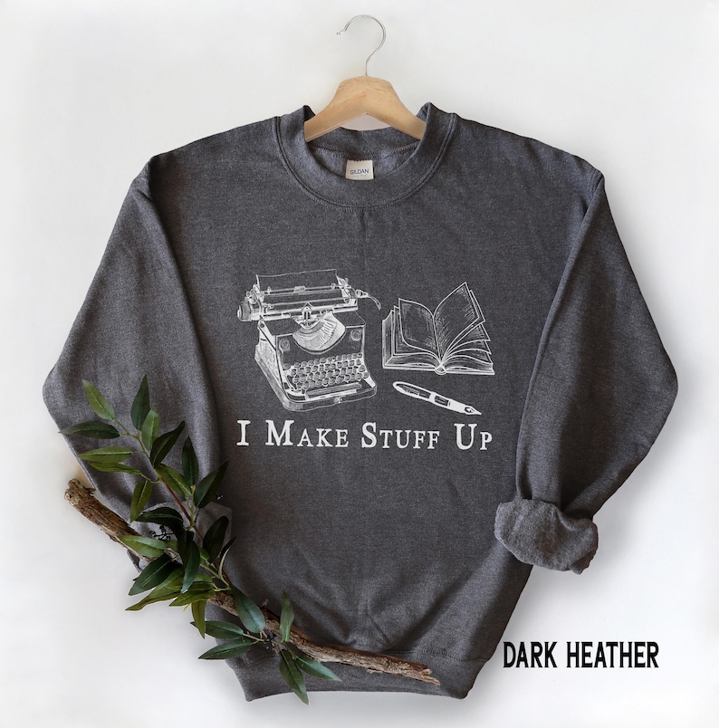 Puede incluir: Sudadera gris oscuro con un gr&aacute;fico blanco de una m&aacute;quina de escribir, un libro abierto y un bol&iacute;grafo. El texto "I MAKE STUFF UP" est&aacute; impreso debajo. Las palabras "DARK HEATHER" est&aacute;n impresas en la parte inferior derecha.