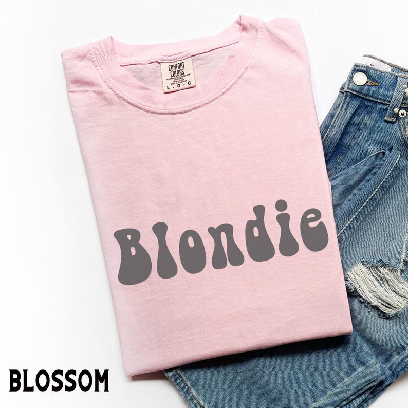 Puede incluir: Una camiseta rosa claro con la palabra "Blondie" en letras grises de estilo retro. La camiseta se muestra con pantalones cortos de mezclilla. La etiqueta de la marca "Comfort Colors" es visible en el cuello de la camiseta.