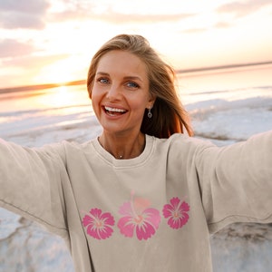 May include: A woman wearing a light beige sweatshirt with a pink hibiscus flower design. She is smiling and taking a selfie at the beach.