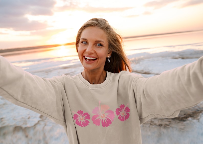 May include: A woman wearing a light beige sweatshirt with a pink hibiscus flower design. She is smiling and taking a selfie at the beach.