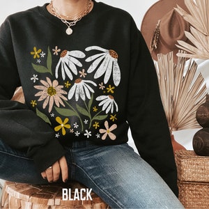 K&ouml;nnte beinhalten: Schwarzer Pullover mit einem Blumendruck mit wei&szlig;en, rosa und gelben Blumen auf schwarzem Hintergrund. Der Pullover hat einen Rundhalsausschnitt und lange &Auml;rmel.
