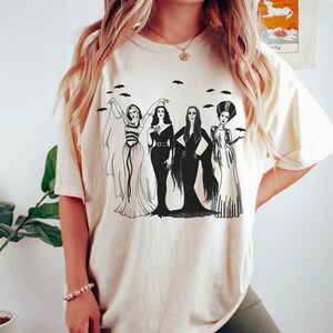 K&ouml;nnte beinhalten: Ein wei&szlig;es T-Shirt mit einem schwarz-wei&szlig;en Grafikdruck von vier Frauen in Vintage-Kleidern. Die Frauen stehen in einer Reihe und haben langes Haar. Die Grafik enth&auml;lt Flederm&auml;use, die &uuml;ber den Frauen fliegen.