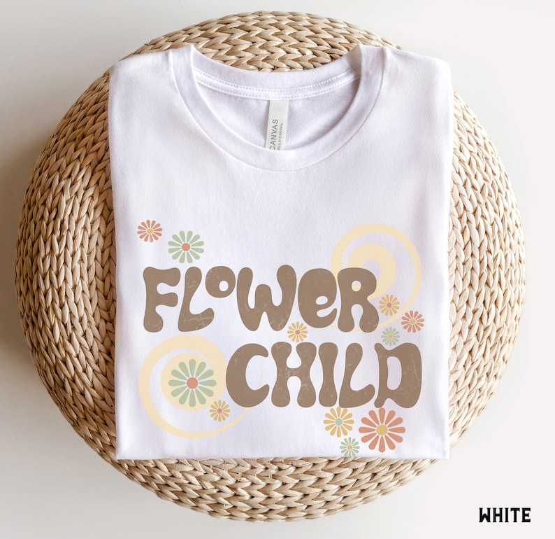 Puede incluir: Una camiseta blanca con las palabras "FLOWER CHILD" en marr&oacute;n, con letras de estilo retro. El dise&ntilde;o incluye flores y espirales en colores pastel. La camiseta est&aacute; doblada cuidadosamente sobre una superficie circular tejida.
