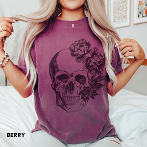 May include: Berry-colored t-shirt featuring a detailed skull graphic adorned with floral elements. The design is printed in dark tones, creating a striking contrast against the shirt's hue. The word "BERRY" is printed at the bottom of the shirt.
