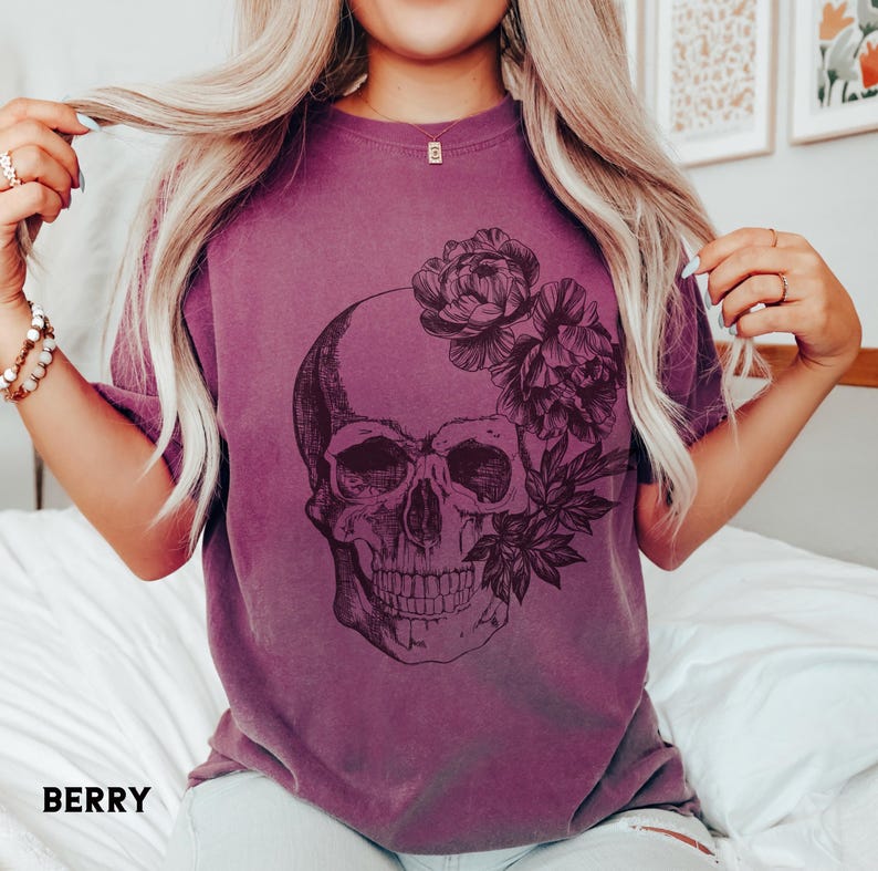 May include: Berry-colored t-shirt featuring a detailed skull graphic adorned with floral elements. The design is printed in dark tones, creating a striking contrast against the shirt's hue. The word "BERRY" is printed at the bottom of the shirt.