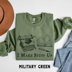 Puede incluir: Sudadera verde militar con un gr&aacute;fico negro de una m&aacute;quina de escribir, un libro abierto y un bol&iacute;grafo. El texto "I Make Stuff Up" est&aacute; debajo del gr&aacute;fico. Las palabras "MILITARY GREEN" est&aacute;n en la parte inferior de la imagen.