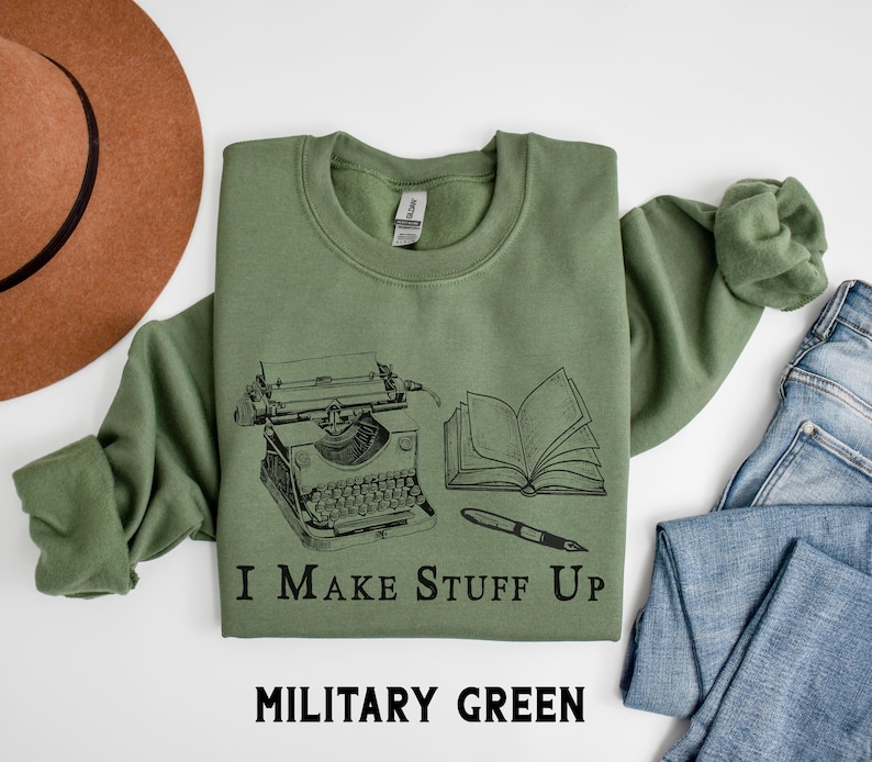 Puede incluir: Sudadera verde militar con un gr&aacute;fico negro de una m&aacute;quina de escribir, un libro abierto y un bol&iacute;grafo. El texto "I Make Stuff Up" est&aacute; debajo del gr&aacute;fico. Las palabras "MILITARY GREEN" est&aacute;n en la parte inferior de la imagen.