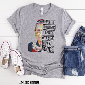 Op de afbeelding: Athletic heather grijze t-shirt met een kleurrijke afbeelding van een vrouwen gezicht en de tekst "NEVER UNDERESTIMATE THE POWER OF A GIRL WITH A BOOK." Het shirt wordt gecombineerd met een blauwe jeans en witte sneakers.