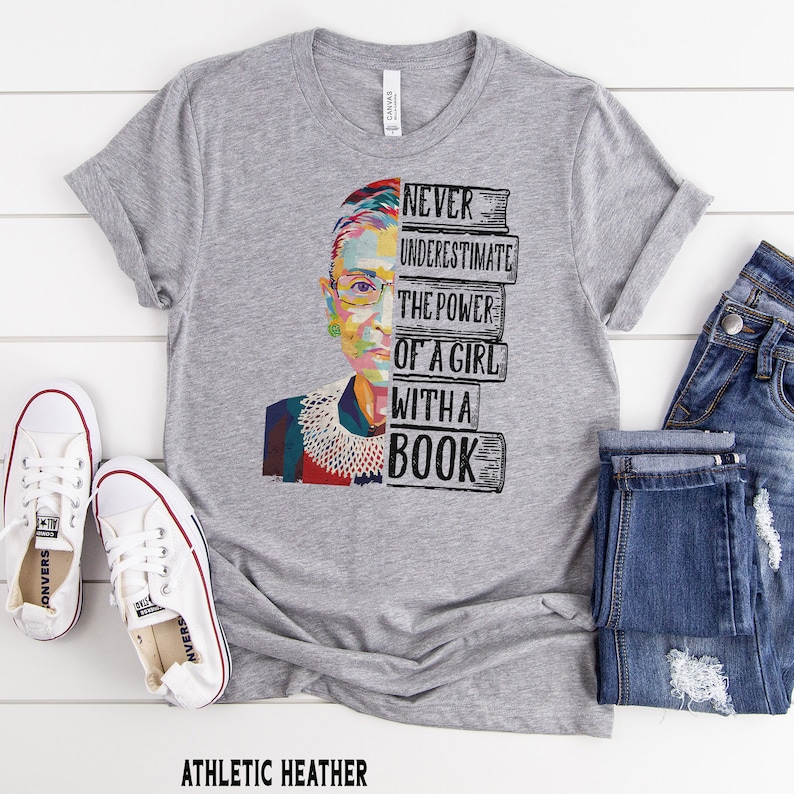 Op de afbeelding: Athletic heather grijze t-shirt met een kleurrijke afbeelding van een vrouwen gezicht en de tekst "NEVER UNDERESTIMATE THE POWER OF A GIRL WITH A BOOK." Het shirt wordt gecombineerd met een blauwe jeans en witte sneakers.