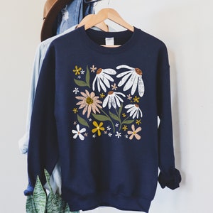 K&ouml;nnte beinhalten: Ein marineblauer Pullover mit einem Blumendruck mit wei&szlig;en, rosa, gelben und gr&uuml;nen Blumen. Die Blumen sind stilisiert und haben einen Vintage-Look.