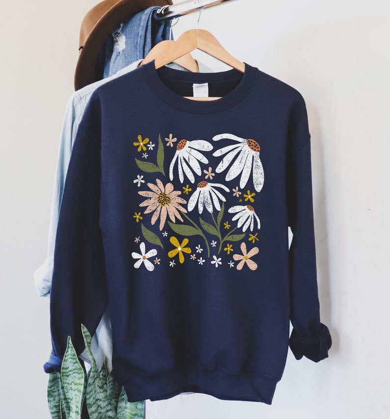 K&ouml;nnte beinhalten: Ein marineblauer Pullover mit einem Blumendruck mit wei&szlig;en, rosa, gelben und gr&uuml;nen Blumen. Die Blumen sind stilisiert und haben einen Vintage-Look.