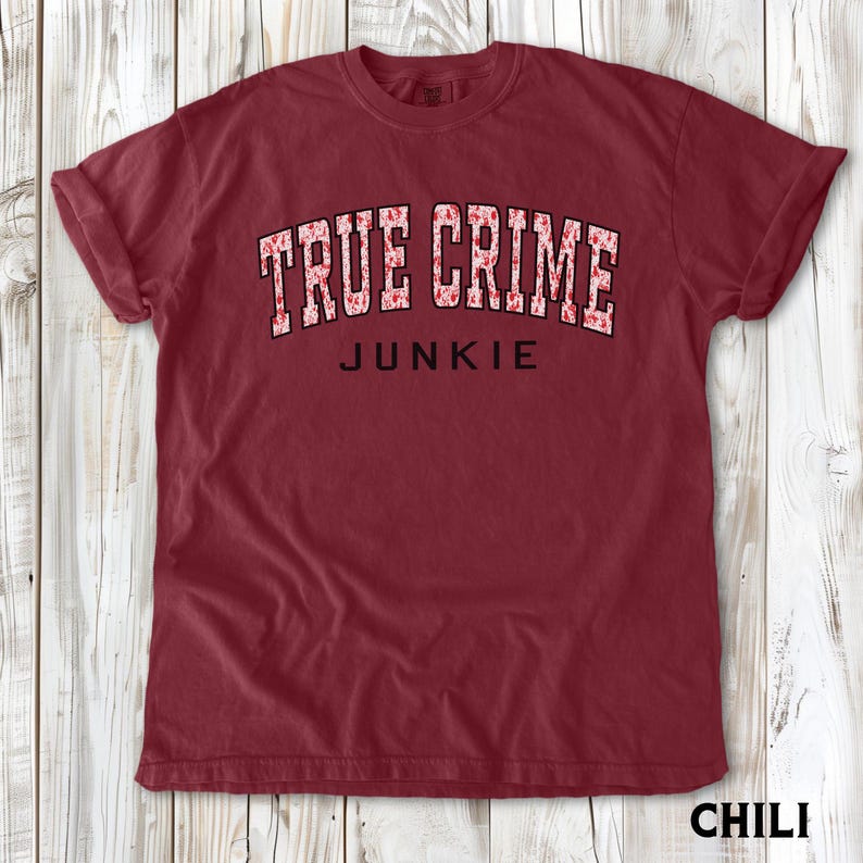 K&ouml;nnte beinhalten: Ein weinrotes T-Shirt mit hochgekrempelten &Auml;rmeln. Das Shirt zeigt den Text "TRUE CRIME JUNKIE" in einer wei&szlig;en, floral gemusterten Schriftart. Das Wort "JUNKIE" ist kleiner und schwarz. Das Wort "CHILI" steht unten.