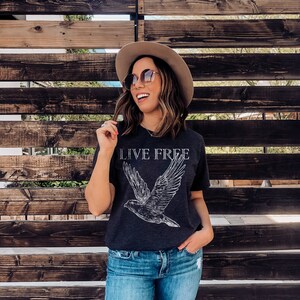 Op de afbeelding: Een vrouw draagt een donkergrijs T-shirt met een witte vogelprint en de tekst "LIVE FREE". Ze draagt ook een bruine hoed, een zonnebril en een blauwe jeans.