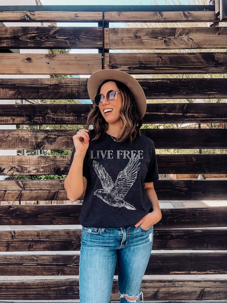 Op de afbeelding: Een vrouw draagt een donkergrijs T-shirt met een witte vogelprint en de tekst "LIVE FREE". Ze draagt ook een bruine hoed, een zonnebril en een blauwe jeans.