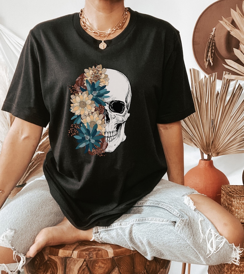Op de afbeelding: Een zwart T-shirt met een afbeelding van een schedel met bloemen die de ene kant van de schedel bedekken. De bloemen zijn in tinten geel, blauw en bruin.