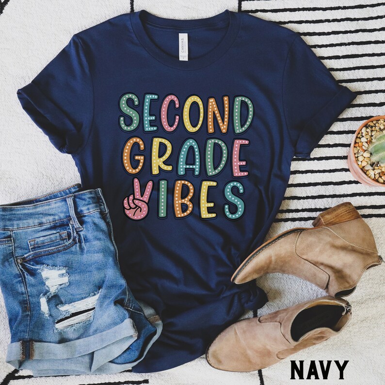 May include: A navy blue t-shirt with the text "Second Grade Vibes" in colorful letters with white polka dots. A peace sign is to the left of the text. The shirt is laying on a white and black striped surface.