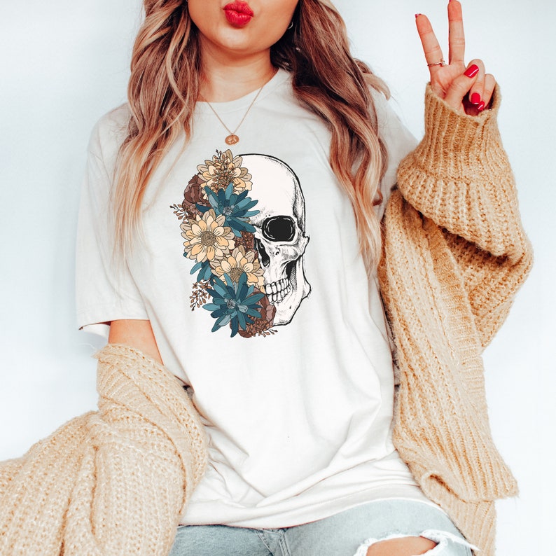 Op de afbeelding: Een wit T-shirt met een afbeelding van een schedel met bloemen aan de ene kant. De bloemen zijn blauw, geel en bruin.