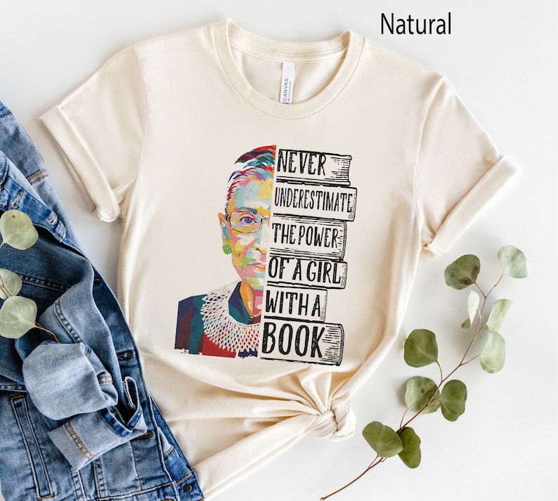 Op de afbeelding: Cr&egrave;mekleurig T-shirt met een kleurrijk portret van een vrouw met een bril en een witte kraag. De tekst "NEVER UNDERESTIMATE THE POWER OF A GIRL WITH A BOOK" staat rechts op de afbeelding.