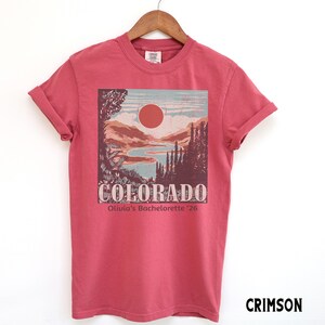 May include: Crimson-colored t-shirt featuring a Colorado landscape design with a sun, mountains, and trees. The word "COLORADO" is printed across the bottom of the design, with "Olivia's Bachelorette '26" below it. The shirt is displayed on a wooden hanger.